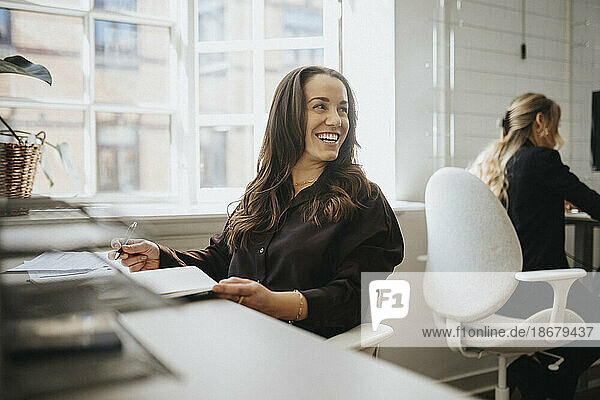 Glückliche Unternehmerin  die wegschaut  während sie am Schreibtisch im Büro sitzt