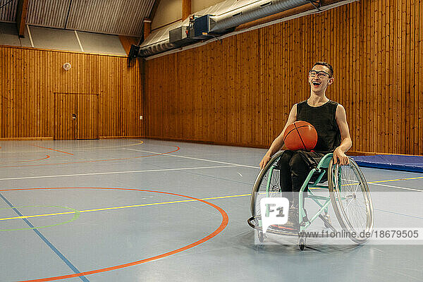 Glückliches Mädchen mit Basketball im Rollstuhl sitzend auf einem Sportplatz
