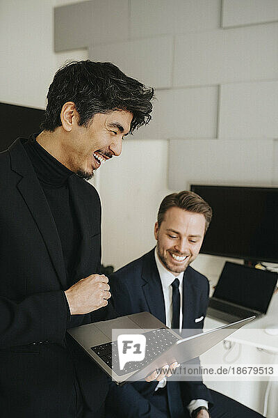Happy businessman holding laptop by male colleague in office