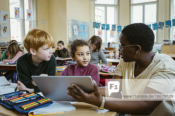 Female teacher assisting schoolboy and schoolgirl with digital tablet in classroom