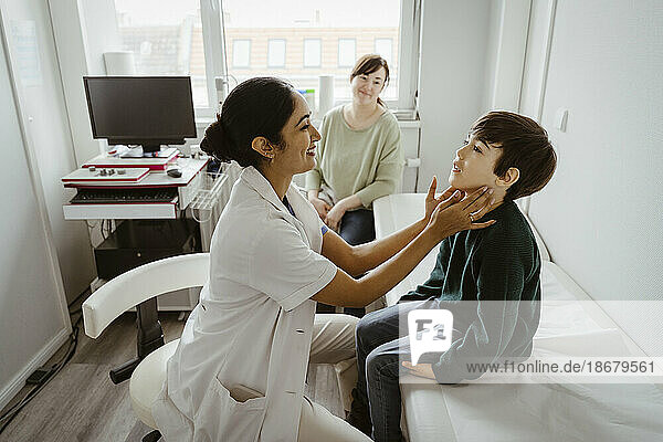 Seitenansicht einer lächelnden Kinderärztin  die einen Jungen untersucht  der auf einem Bett im Untersuchungsraum einer Klinik sitzt