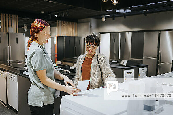 Saleswoman advising female customer in buying washing machine at electronics store