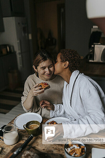 Non-binary person kissing friend while having breakfast at home