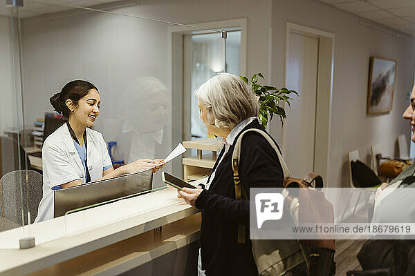 Lächelnde Empfangsdame erklärt dem Patienten das Ausfüllen von Formularen durch einen transparenten Schild in der Klinik