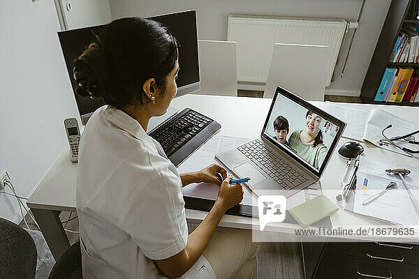 Hochformatige Ansicht einer Kinderärztin  die Mutter und Sohn per Videoanruf auf einem Laptop in einer Klinik berät