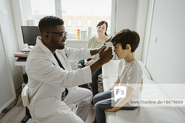 Männlicher Kinderarzt  der einen Jungen untersucht  der auf einem Bett im Untersuchungsraum eines Gesundheitszentrums sitzt