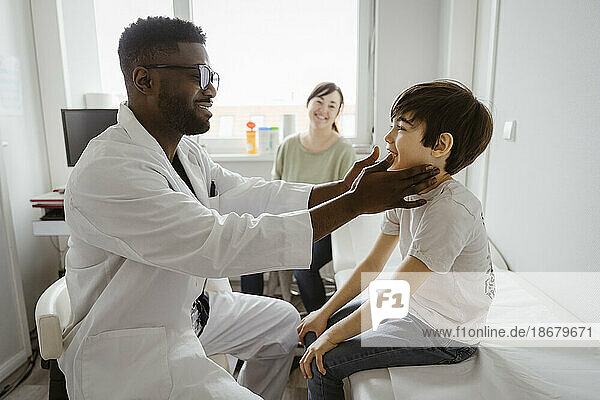 Lächelnder männlicher Kinderarzt  der den Hals eines Jungen untersucht  der in einem Gesundheitszentrum auf einem Bett sitzt
