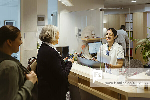 Patients talking to female receptionist through transparent shield in clinic