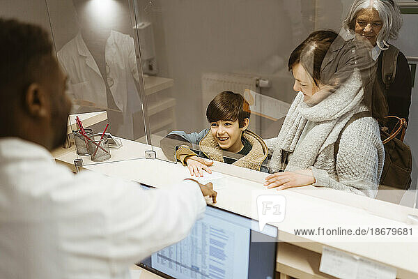 Lächelnder Junge reicht lächelnd ein Dokument an einen männlichen Rezeptionisten unter einem transparenten Schild in einer Klinik