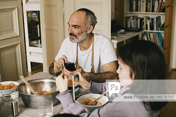 Father and son sitting with smart phones and bowls at dining table