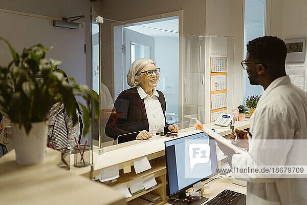 Lächelnde ältere Patientin im Gespräch mit einem männlichen Rezeptionisten durch einen transparenten Schild in einer Klinik