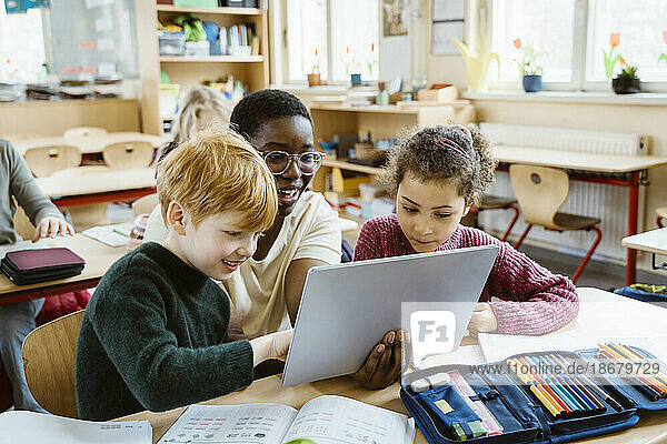 Glücklicher Lehrer  der einem Jungen und einem Mädchen mit einem digitalen Tablet hilft  die am Schreibtisch im Klassenzimmer sitzen