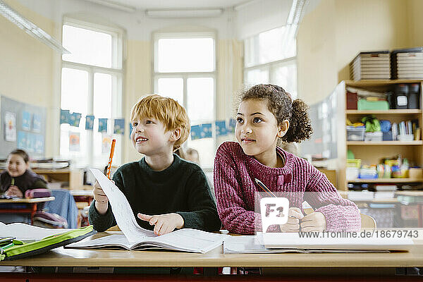 Smiling blond boy sitting with girl at desk looking away in classroom