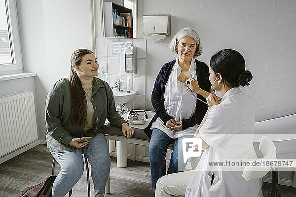 Doctor listening heartbeat of senior woman sitting with daughter in clinic