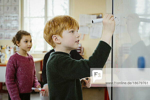 Seitenansicht eines blonden Jungen  der auf eine Tafel schreibt  während er eine mathematische Aufgabe im Klassenzimmer löst