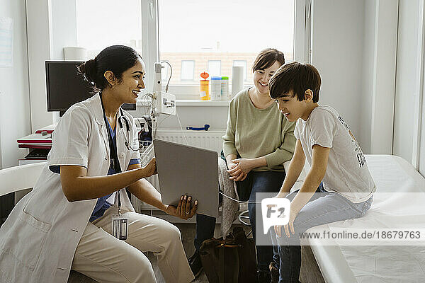 Eine Kinderärztin zeigt einer Mutter und ihrem Sohn  die im Untersuchungsraum einer Klinik sitzen  einen Tablet-PC