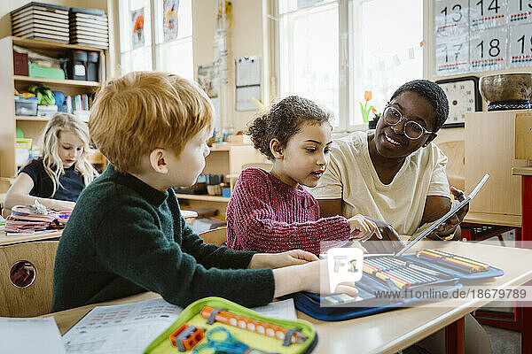 Lächelnder Lehrer  der einem Jungen und einem Mädchen mit einem digitalen Tablet hilft  die am Schreibtisch im Klassenzimmer sitzen