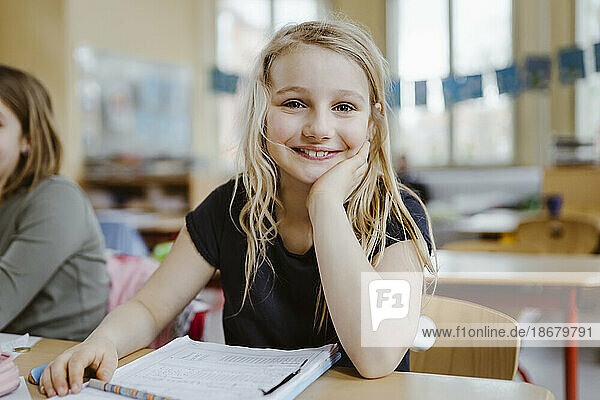 Smiling blond schoolgirl leaning on elbow while sitting at desk in classroom