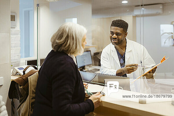 Lächelnder männlicher Empfangsmitarbeiter  der ein Klemmbrett hält  während er durch einen transparenten Schild mit einer älteren Frau in einer Klinik spricht