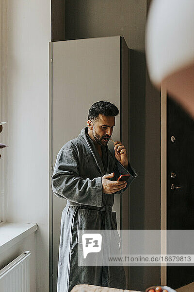 Mann im Bademantel benutzt Smartphone beim Zähneputzen zu Hause