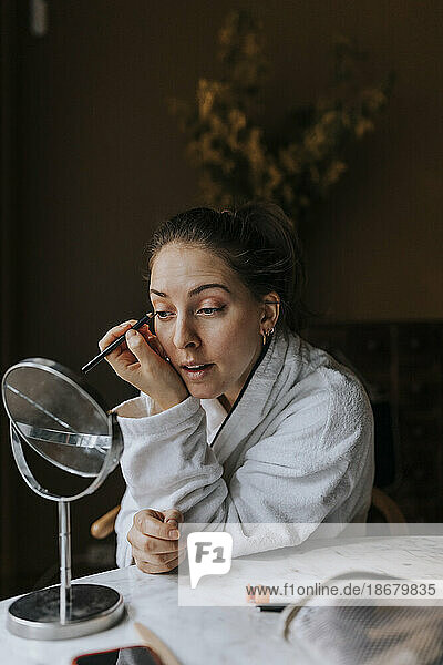 Nicht-binäre Person trägt Make-up auf  während sie sich zu Hause im Spiegel betrachtet