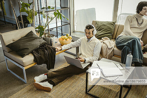 High angle view of programmer using mobile phone sitting with laptop on carpet in creative office