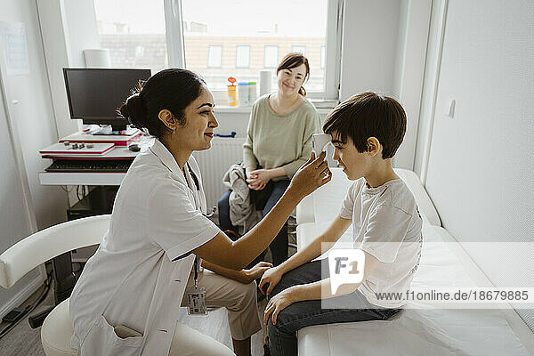 Smiling female pediatrician measuring body temperature of boy through infrared thermometer at clinic