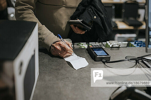 Mittelteil eines älteren Mannes  der eine Rechnung mit einem Kreditkartenleser an der Kasse eines Elektronikgeschäfts unterschreibt
