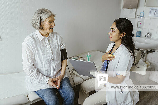 Female doctor holding tablet PC advising senior patient sitting on bed in clinic