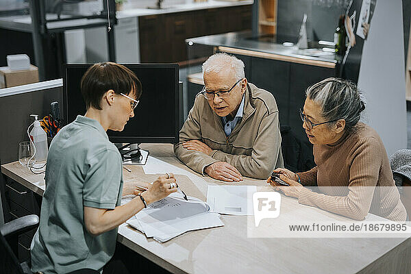 Eine Innenarchitektin diskutiert mit einem älteren Ehepaar an einem Schreibtisch in einem Geschäft über eine Broschüre