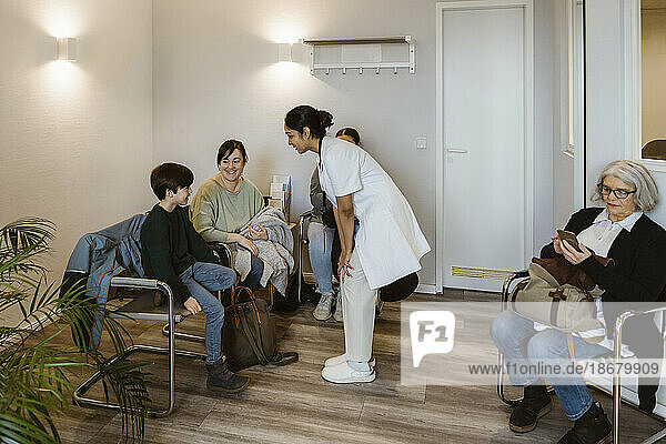 Ärztin mit Händen auf dem Knie im Gespräch mit einem Jungen  der mit seiner Mutter im Wartezimmer sitzt