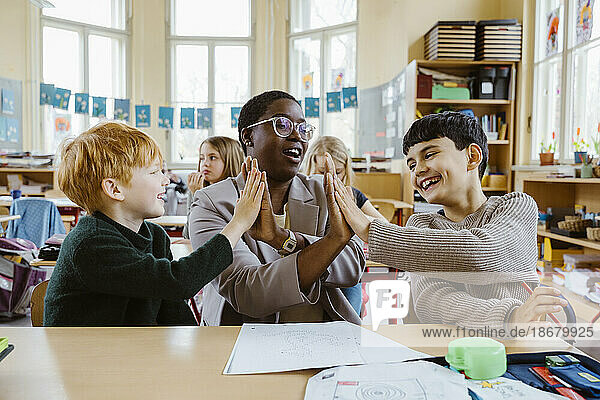 Glückliche Lehrerin und Jungen geben sich High-Fives  während sie am Schreibtisch im Klassenzimmer sitzen