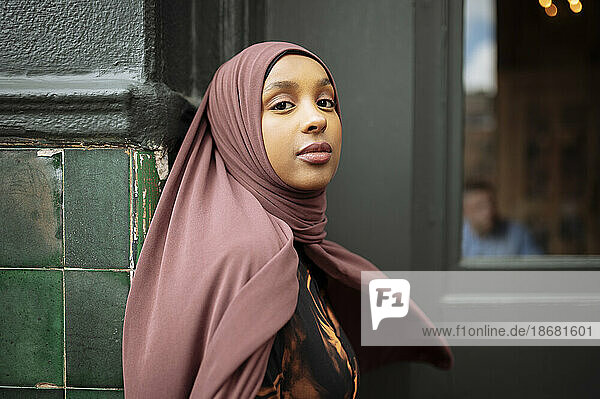 Portrait of young woman in hijab leaning against building
