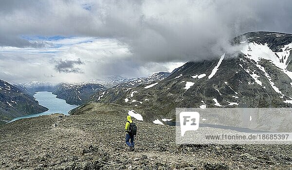 Bergsteiger auf steinigem Wanderweg  Besseggen Wanderung  Gratwanderung  Ausblick auf See Gjende und verschneite Berge  Jotunheimen Nationalpark  Vågå  Innlandet  Norwegen  Europa