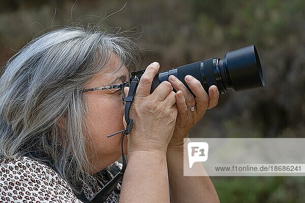 Older woman with white hair and glasses taking a photo looking through the viewfinder of her camera with trees in the background