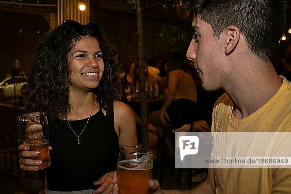 Junge lateinamerikanische Frau lächelt und plaudert mit einem jungen Mann in einer Bar und trinkt Bier. Treffen bei einem Online Date. Unscharfer Hintergrund