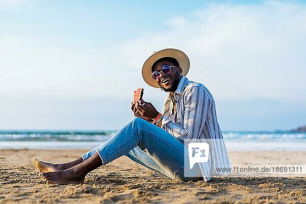 Portrait of a black ethnic man enjoy summer vacation at the beach eating a watermelon  Sitting on the sand smiling