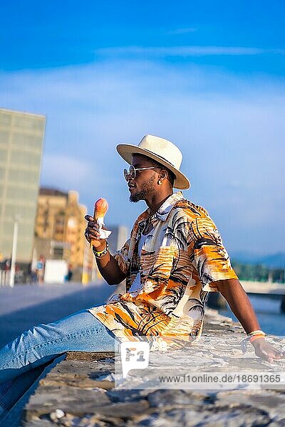 Porträt eines schwarzen ethnischen Mannes  der den Sommerurlaub am Strand genießt  ein Eis isst und am Fluss sitzt