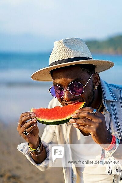 Black ethnic man enjoy summer vacation on the beach eating a watermelon