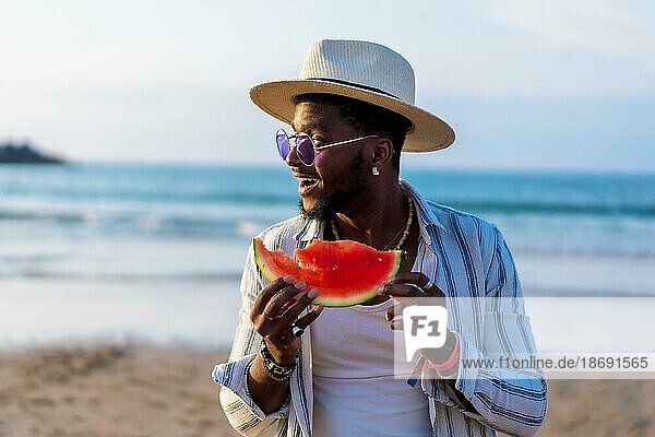 Black ethnic man enjoy summer vacation on the beach eating a watermelon by the sea