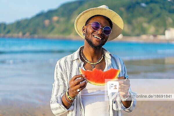 Black ethnic man enjoy summer vacation at the beach eating a watermelon smiling