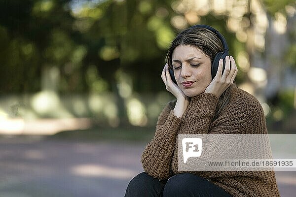Junge Frau sitzt mit nachdenklichem Gesichtsausdruck in einem Park und hört Musik