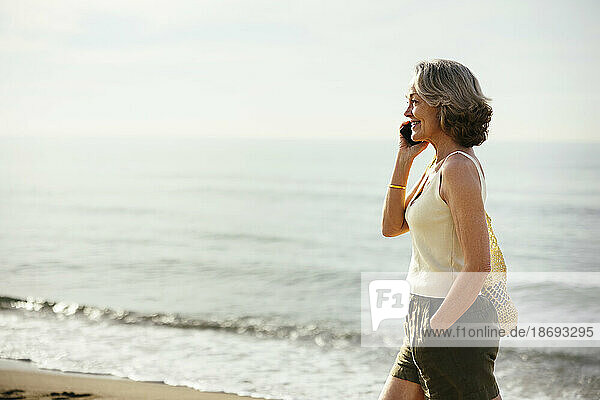 Lächelnde Frau mit der Hand in der Tasche  die am Strand mit dem Smartphone spricht