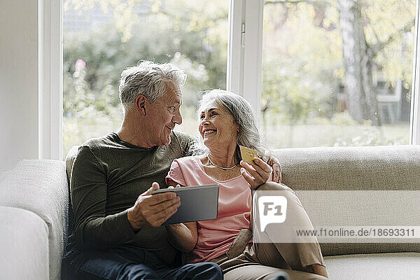 Glückliches Seniorenpaar entspannt sich zu Hause auf der Couch und nutzt das Tablet zum Online-Shopping