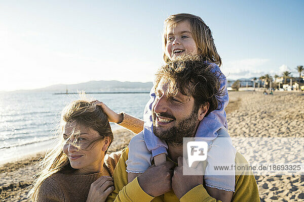 Smiling man with daughter sitting on shoulders by woman at beach