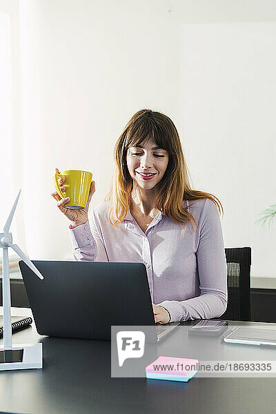 Smiling businesswoman holding coffee cup and working on laptop in office