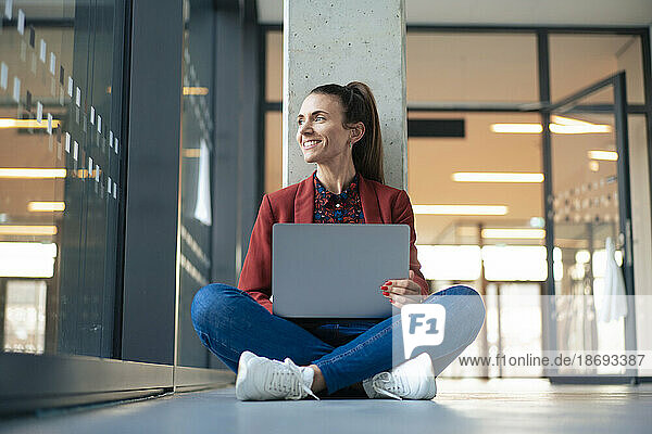 Lächelnde Geschäftsfrau mit Laptop sitzt im Schneidersitz auf dem Boden im Büro