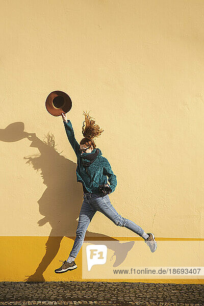 Woman jumping with hat and camera in front of yellow wall