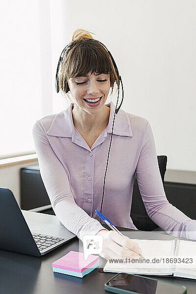 Happy businesswoman wearing headset writing on note pad at desk