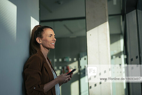 Nachdenkliche Geschäftsfrau hält Smartphone im Büro an die Wand gelehnt